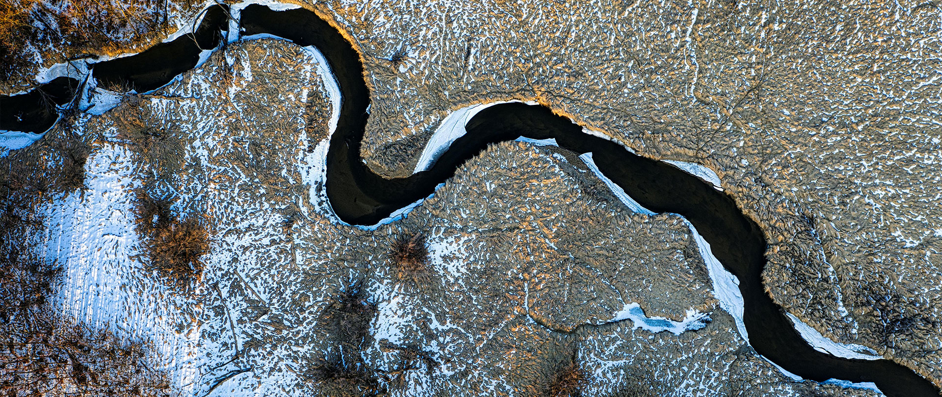 Aerial view of a winding river through a landscape of snow and earth, showcasing a contrast of textures and colors in the winter environment.