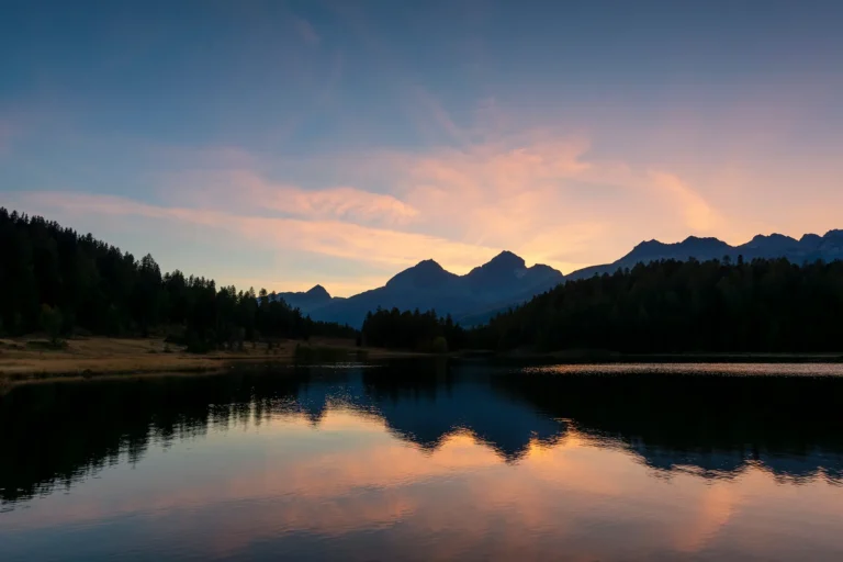 Siluetas de montañas y bosque reflejados en un lago tranquilo bajo un cielo de puesta de sol lleno de color.