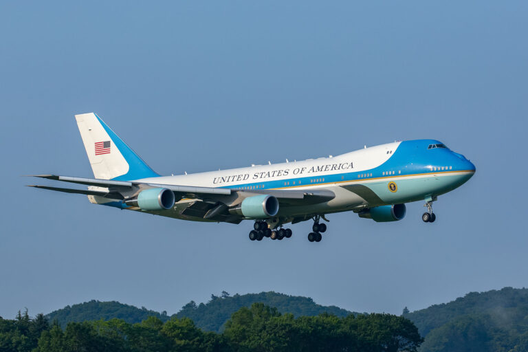 Air Force One en train de descendre pour atterrir dans un ciel bleu clair, au-dessus de collines boisées.