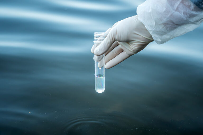 Gloved hand collecting a water sample in a test tube from a natural water source for PFAS contamination testing.